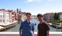 two men standing on a bridge over a river with buildings in the background